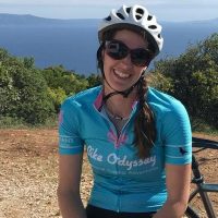 Isabel Hofman Isabel Hofman, Bike Odyssey tour leader, smiling in helmet and sunglasses beside her bike overlooking the coast