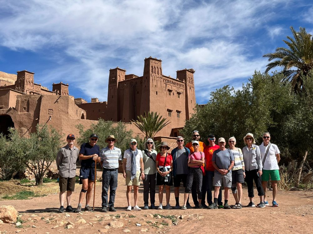 A group of cyclists and walkers posing together in front of Ait Benhaddou's kasbah towers.