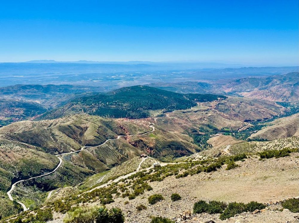 A sweeping view of a switchback mountain road winding down through green and red terraced hillsides.