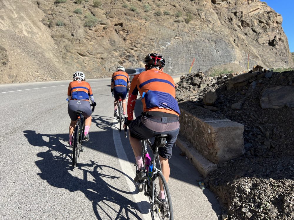 A line of cyclists riding away up a mountain pass road cut into a rocky hillside.