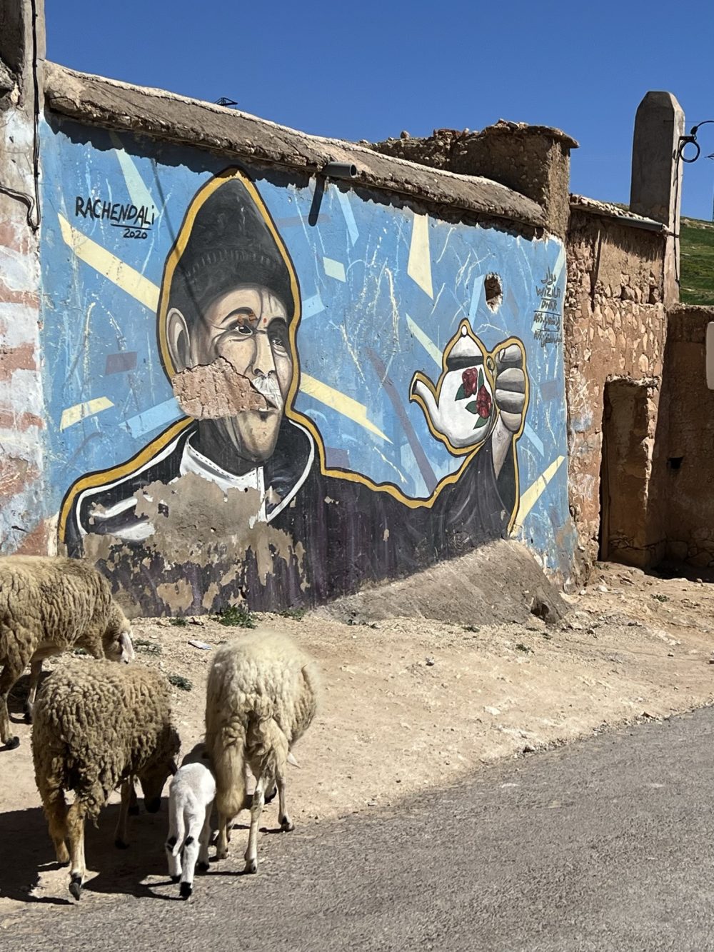 A street-art mural of a monk-like figure holding a teapot on a rural wall, with a small flock of sheep grazing below.