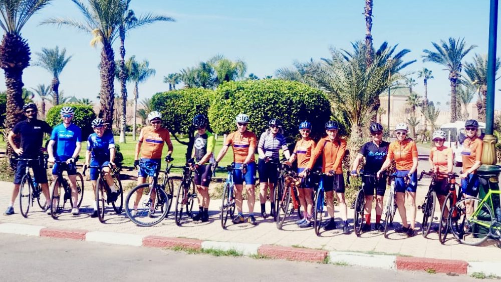 A line-up of cyclists in matching kit posing with their bikes beside palm trees on a Marrakech street.