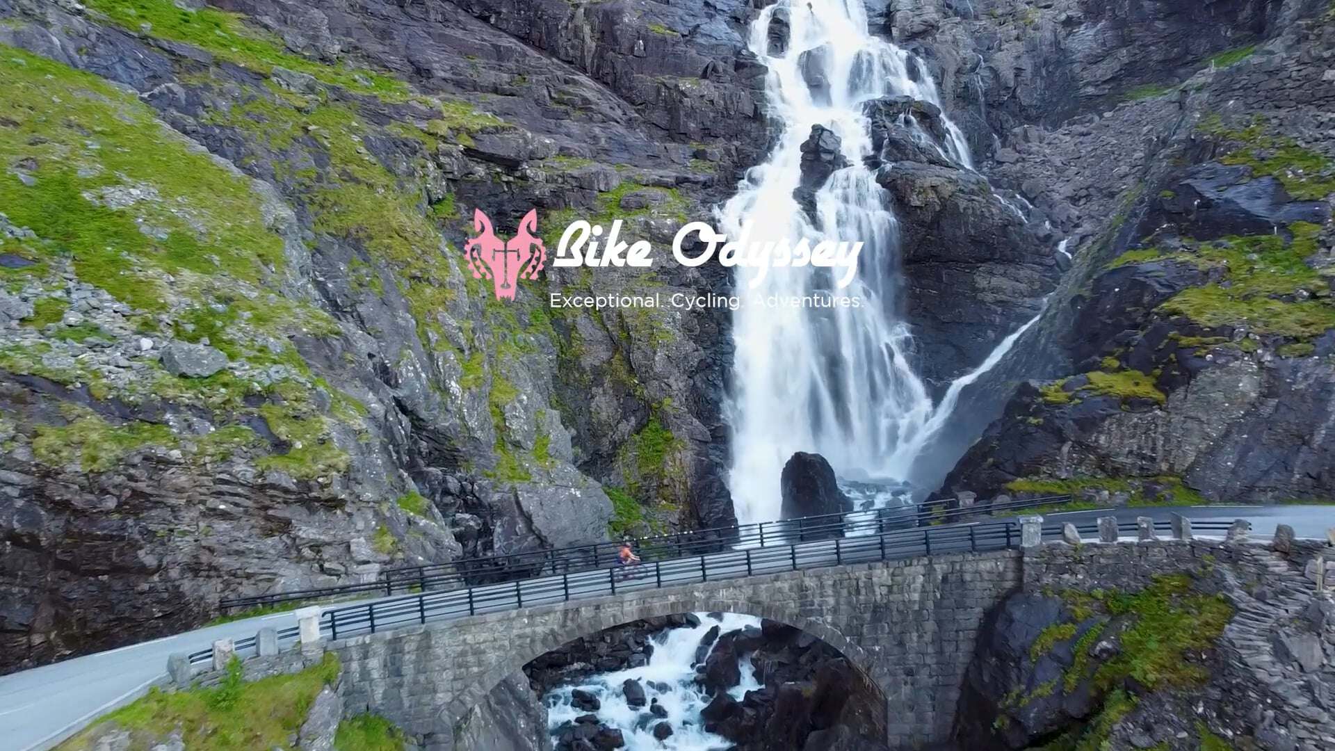 Cyclist crossing a stone arch bridge beneath a mountain waterfall, with the Bike Odyssey logo