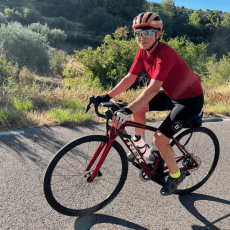 Bike Odyssey guest Peggy O'Neil in a red jersey, smiling on her road bike on a sunny country road on tour.